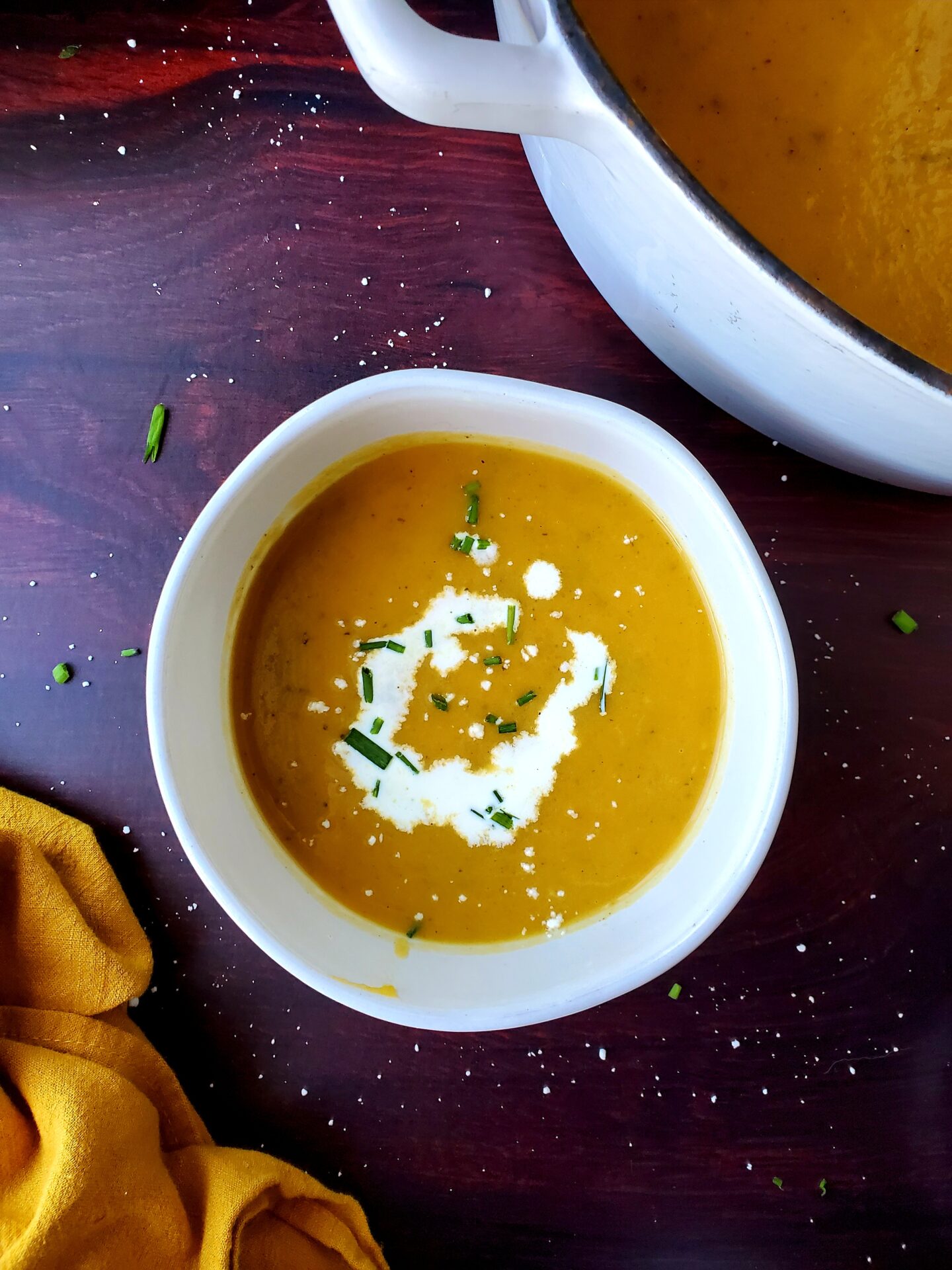 bowl of apple butternut squash soup with swirl of cream; yellow napkin; edge of bowl; replica surfaces mahogany background